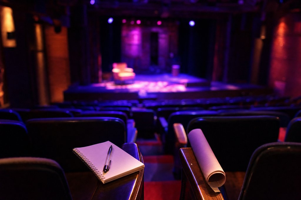 The back rows of a small theatre before curtain, a spiral notebook and pen resting on one seat and a rolled programme on the next, empty rows sloping away toward a dimly lit stage.